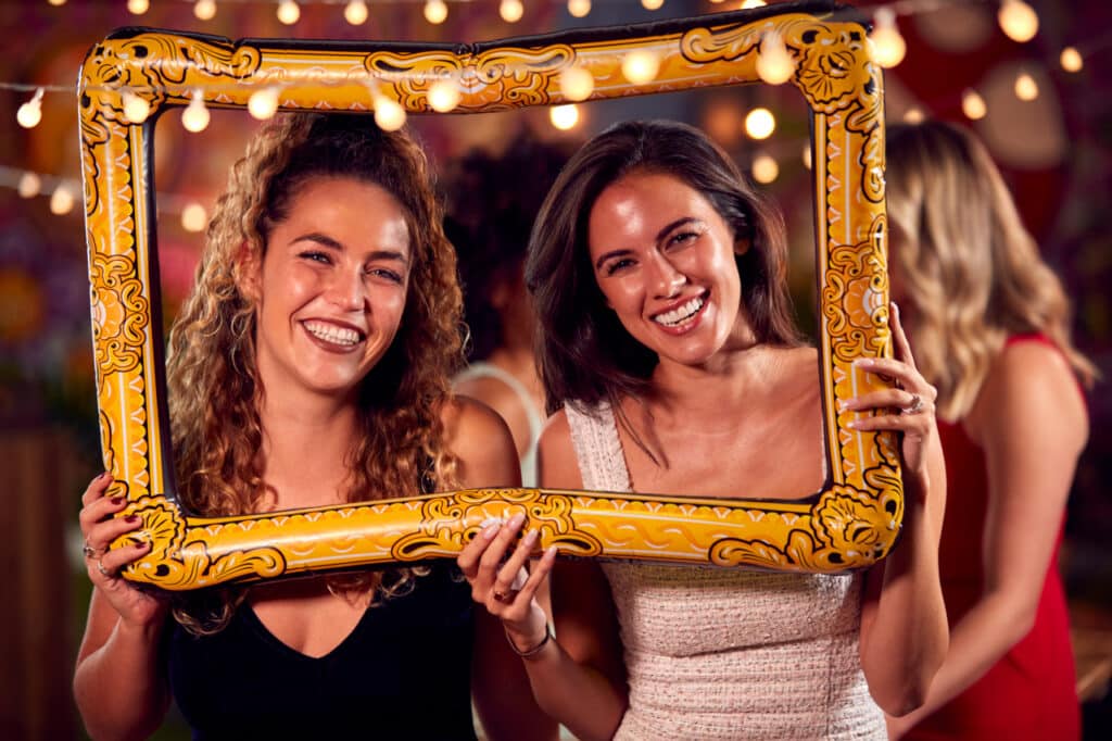 Female Friends Having Fun Posing With Photo Booth Photo Frame At Wedding Party